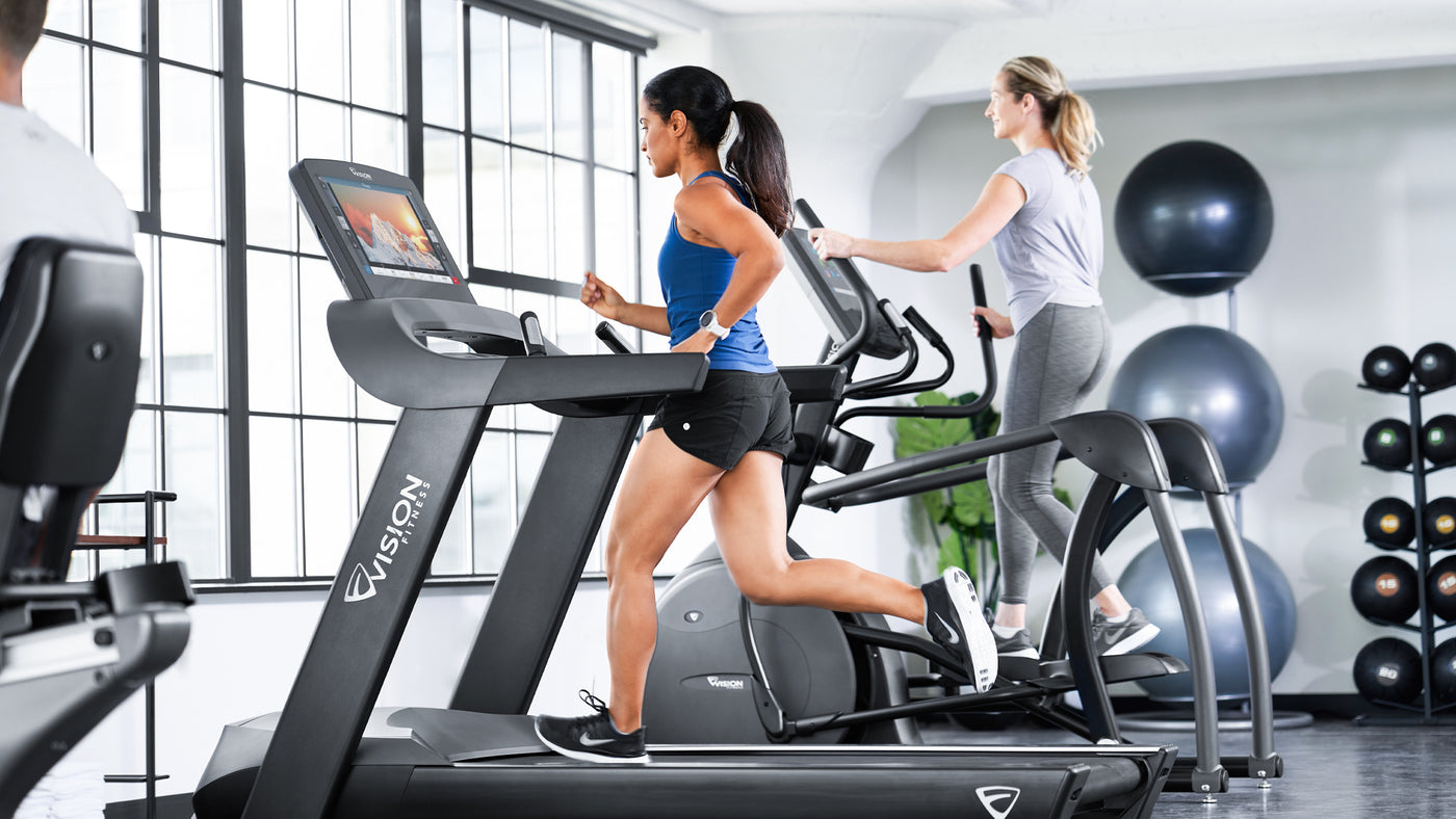 Lady in Short and Blue Top running on Vision Fitness Treadmill in Gym Space - lifestyle image - woman on elliptical background